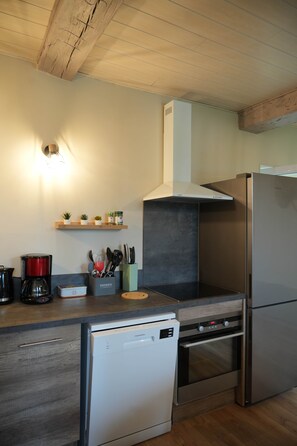 A compact kitchen with a dishwasher, oven, and a built-in stove. There are shelves with kitchen utensils and a coffee maker. - Le Rissas, gite for 4 _ Lacets Ventoux (Beaumont-du-Ventoux)