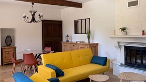 A living room with a yellow sofa, blue armchair, wooden beams, a chandelier, a fireplace, and a dining area. - Le Ventoux, gîte for 5 _ Lacets du Ventoux (Beaumont-du-Ventoux)
