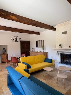 Ein Wohnzimmer mit gelbem Sofa, blauem Sessel, Holzbalken, einer Kristallleuchter, einem Kamin und einem Essbereich.