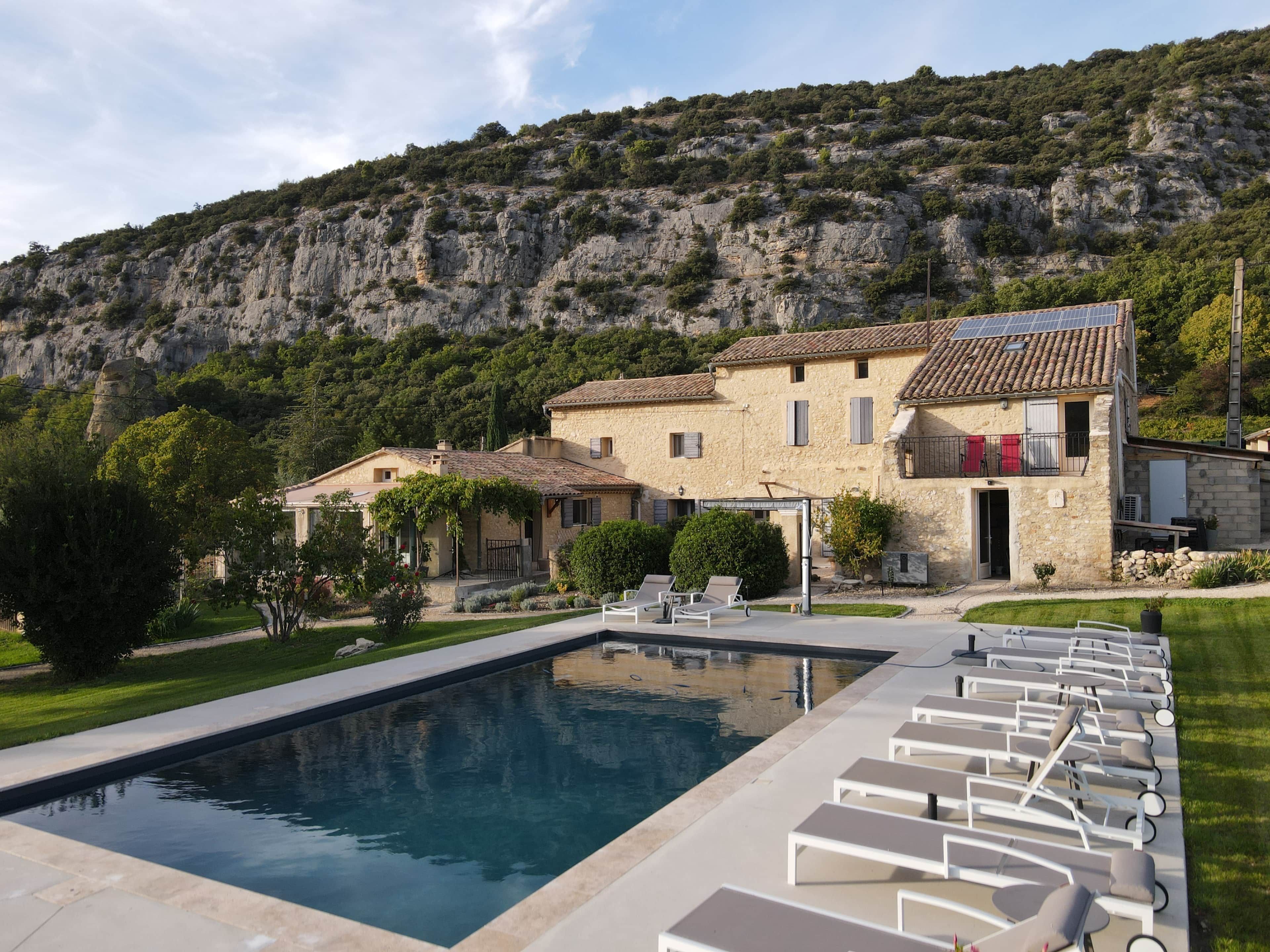 A pool with lounge chairs, a stone building with a tiled roof, and a mountainous backdrop.