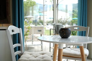 A white chair with a cushioned seat, a round glass table with a dark vase, and a view of outdoor seating and greenery through large windows. - Les Oliviers, gîte for 4p_Lacets du Ventoux (Beaumont-du-Ventoux)