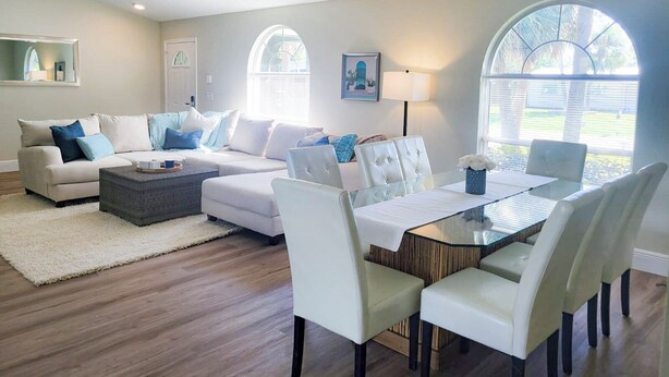 Amplia sala de estar con un juego de sofá color beige, una mesa de centro de vidrio y un comedor con mesa de vidrio y sillas blancas.
