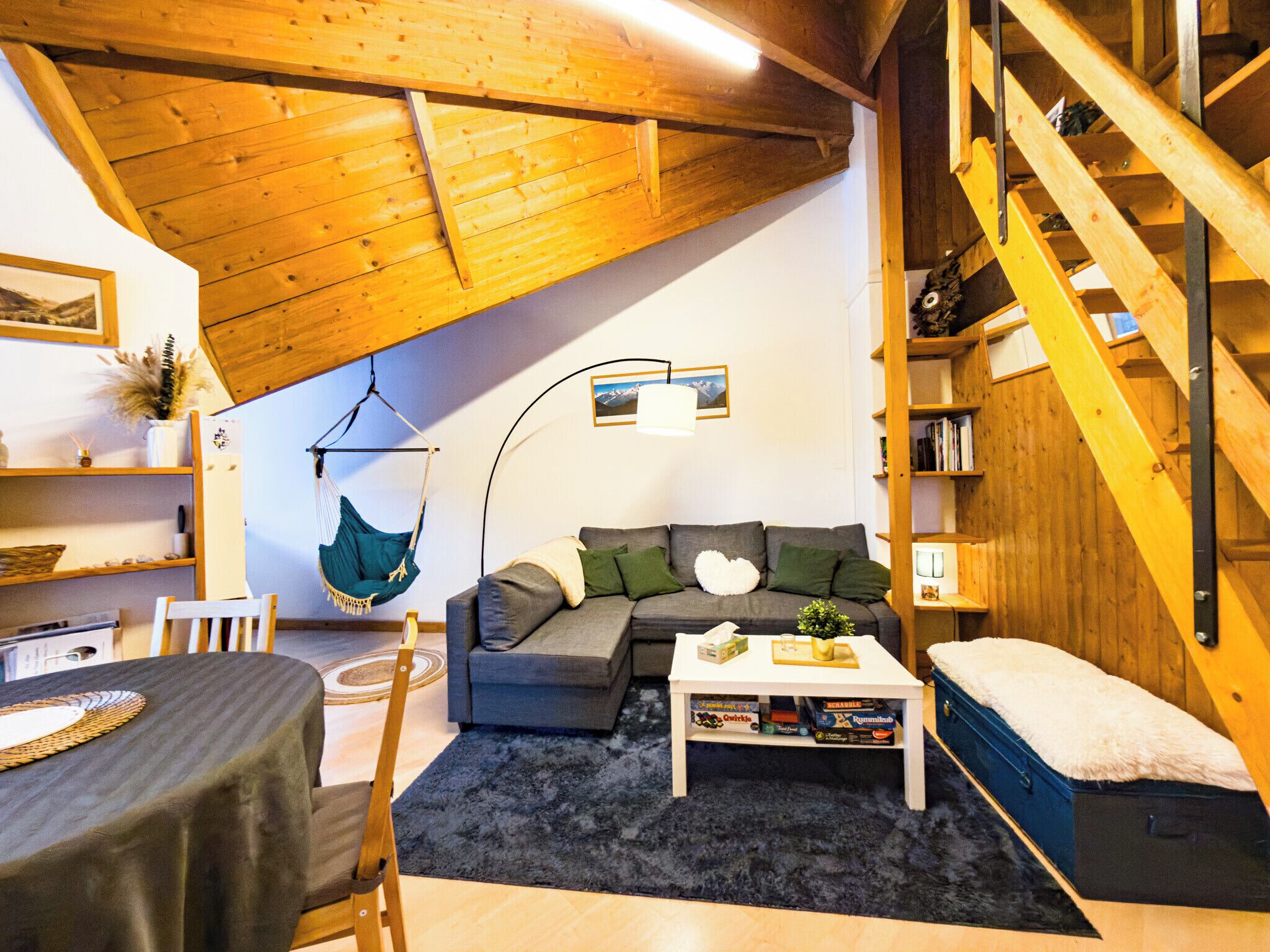 Een gezellige woonkamer met een houten plafond, een hangstoel, een grijze bank, een salontafel en een boekenkast.