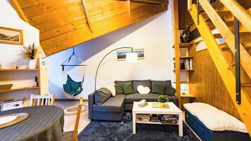 Een gezellige woonkamer met een houten plafond, een hangstoel, een grijze bank, een salontafel en een boekenkast.