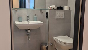 A modern bathroom with a white sink, a glass shower enclosure, a toilet, and a small trash bin. - Green Dream Retreat (Linz)