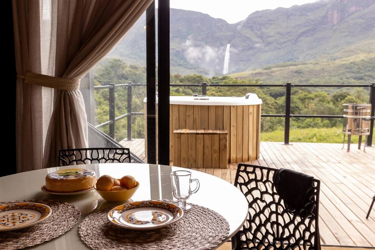 Ein Esstisch mit Tellern, Kuchen und einer Schüssel Brot auf einem Balkon mit Blick auf Berge und einen Wasserfall.