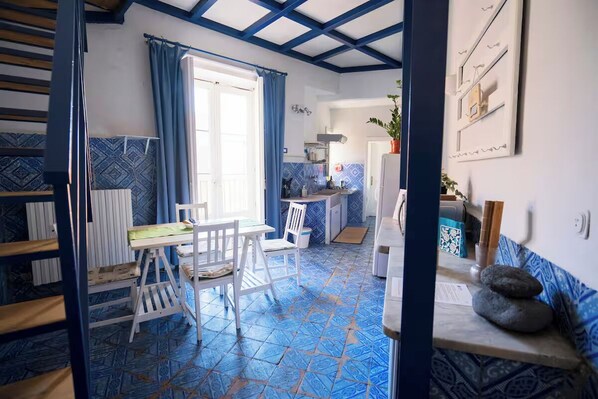 Une pièce avec un sol carrelé de carreaux bleus, une table blanche avec des chaises, une fenêtre avec des rideaux bleus et un coin cuisine équipé d’une cuisinière et de placards.