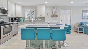A modern kitchen with white cabinets, a central island with bar stools, stainless steel appliances, and a view of the living area with a blue sofa. - 110A Newport Bay ,Deck Seating , Pool , WaterView (Ocean City)