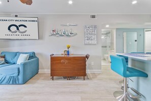 A modern living room with a blue sofa, a wooden dining table, and a kitchen area with blue bar stools. - 110A Newport Bay ,Deck Seating , Pool , WaterView (Ocean City)