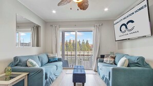 A modern living room with teal sofas, a wooden coffee table, and a large window with a view of trees and buildings. - 110A Newport Bay ,Deck Seating , Pool , WaterView (Ocean City)