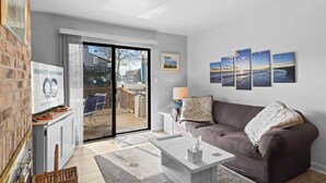 A modern living room with a grey couch, a coffee table, and a TV on a stand. There is a sliding glass door leading to an outdoor area with a deck. - Rustic Harbor I (Ocean City)