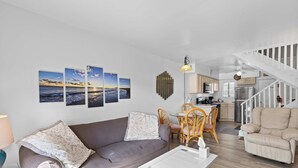 A living room with a sofa, a coffee table, and a dining area with chairs and a table. There is a staircase leading to an upper level. - Rustic Harbor I (Ocean City)