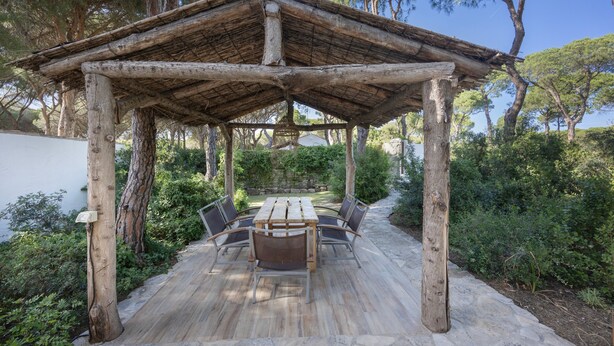 Une pergola en bois avec une table et des chaises, entourée d’un sol en pierre et de verdure.