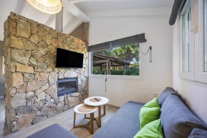 Ein Steinkamin, ein Flachbildfernseher, ein kleiner runder Tisch und ein Sofa mit grünen Kissen.