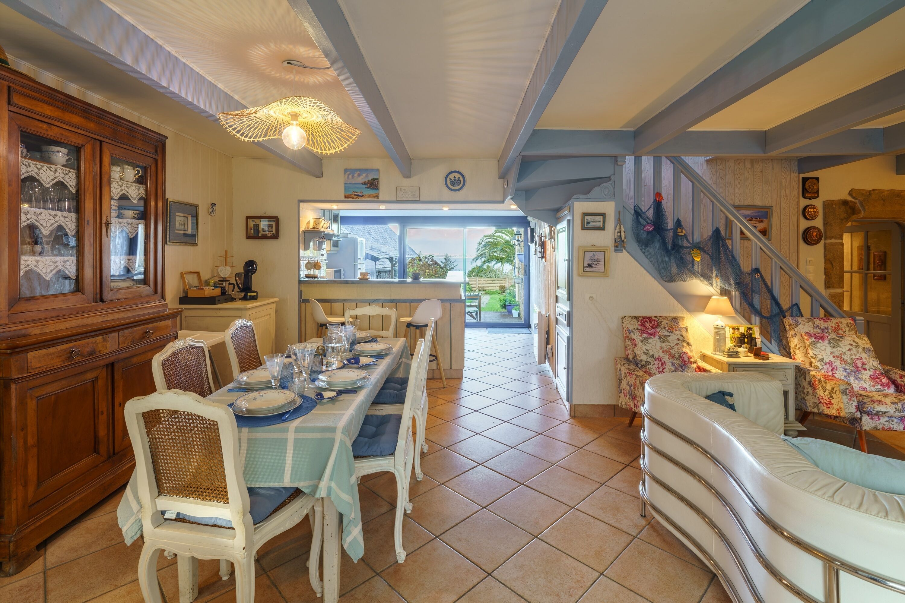 A dining area with a long table set for six, flanked by white chairs with blue cushions. A wooden cabinet with glass doors is to the left, and a staircase with floral upholstery leads to another level.