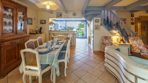 Un'area pranzo con un tavolo lungo apparecchiato per sei, affiancato da sedie bianche con cuscini blu. A sinistra, un mobiletto in legno con ante in vetro, e una scala rivestita di tessuto floreale conduce a un altro livello.