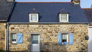 Un edificio in pietra con tetto in ardesia scura, due finestre con persiane blu e una porta bianca.