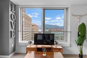 Ein modernes Wohnzimmer mit großem Fenster, einem Flachbildfernseher, einem Holztisch und einer Zimmerpflanze.