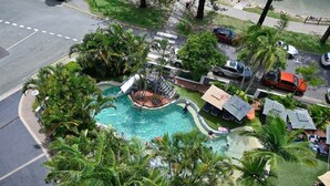 An aerial view of a resort with a central swimming pool surrounded by palm trees and lounge areas with striped umbrellas. - It's Five O'Clock Somewhere (Sunshine Coast)