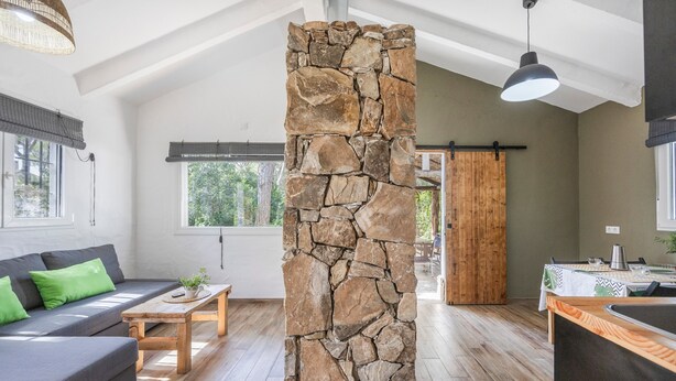 A modern interior with a stone pillar, a sofa with green cushions, a wooden coffee table, and a kitchen area with a table set for dining. - Casa Espartosa 1 in Chiclana with Private Pool, Terrace, and Wi-Fi (Chiclana de la Frontera)