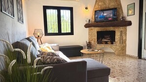A living room with a stone fireplace, a sofa, a television, and a window with bars. - Villa Horizonte with pool and pool house, mountain and nature views (orba)