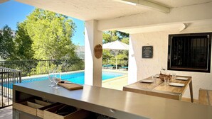 A poolside bar with a view of the pool and trees, a wooden table with glasses and a bottle, and a sign on the wall. - Villa Horizonte with pool and pool house, mountain and nature views (orba)