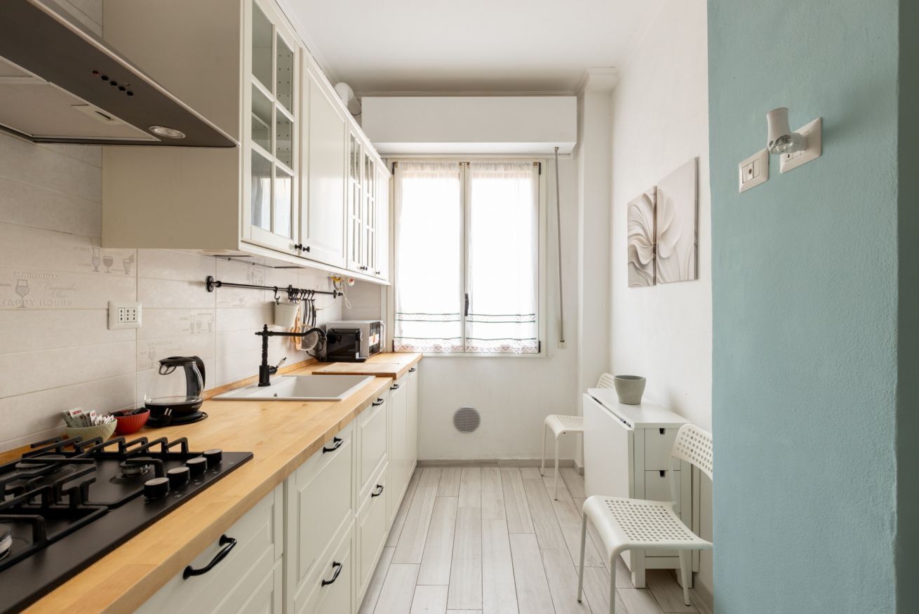 Een kleine keuken met witte kastjes, een houten aanrecht, een gasfornuis, een waterkoker en een klein raam met gordijnen.
