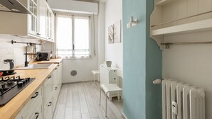 Cocina compacta con estufa, microondas y armarios, una ventana con cortinas, un radiador y un banco blanco.