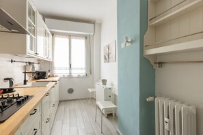 Une cuisine compacte équipée d’une cuisinière, d’un four à micro-ondes et de placards, d’une fenêtre avec des rideaux, d’un radiateur et d’un banc blanc.
