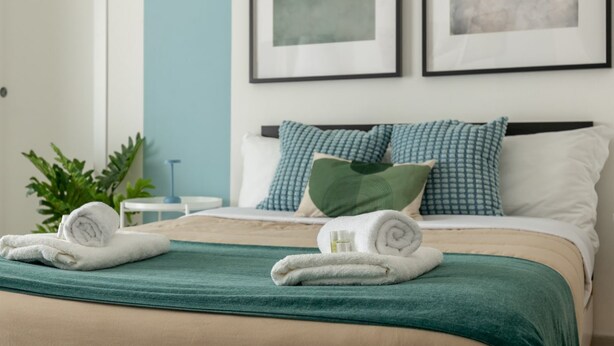 Una cama bien hecha con una manta verde, almohadas blancas y toallas enrolladas.
