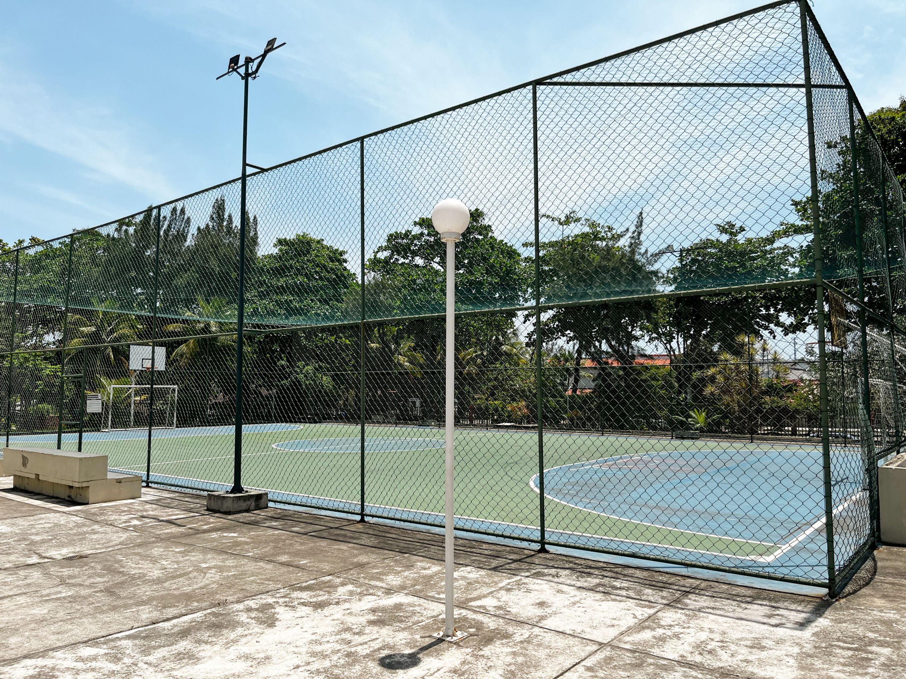 Een tennisbaan omheind door een hek van gaas, met een basketbalveld en een lichtmast.
