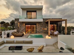 Una moderna casa a due piani con facciata in pietra, un balcone in vetro, una piscina e un'area salotto esterna.