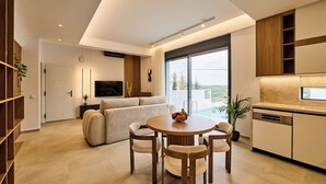 A modern living room with a sofa, dining table, and chairs, a TV, and a large window with a view. - POLYTELIA | Luxury Living (Hersonissos)