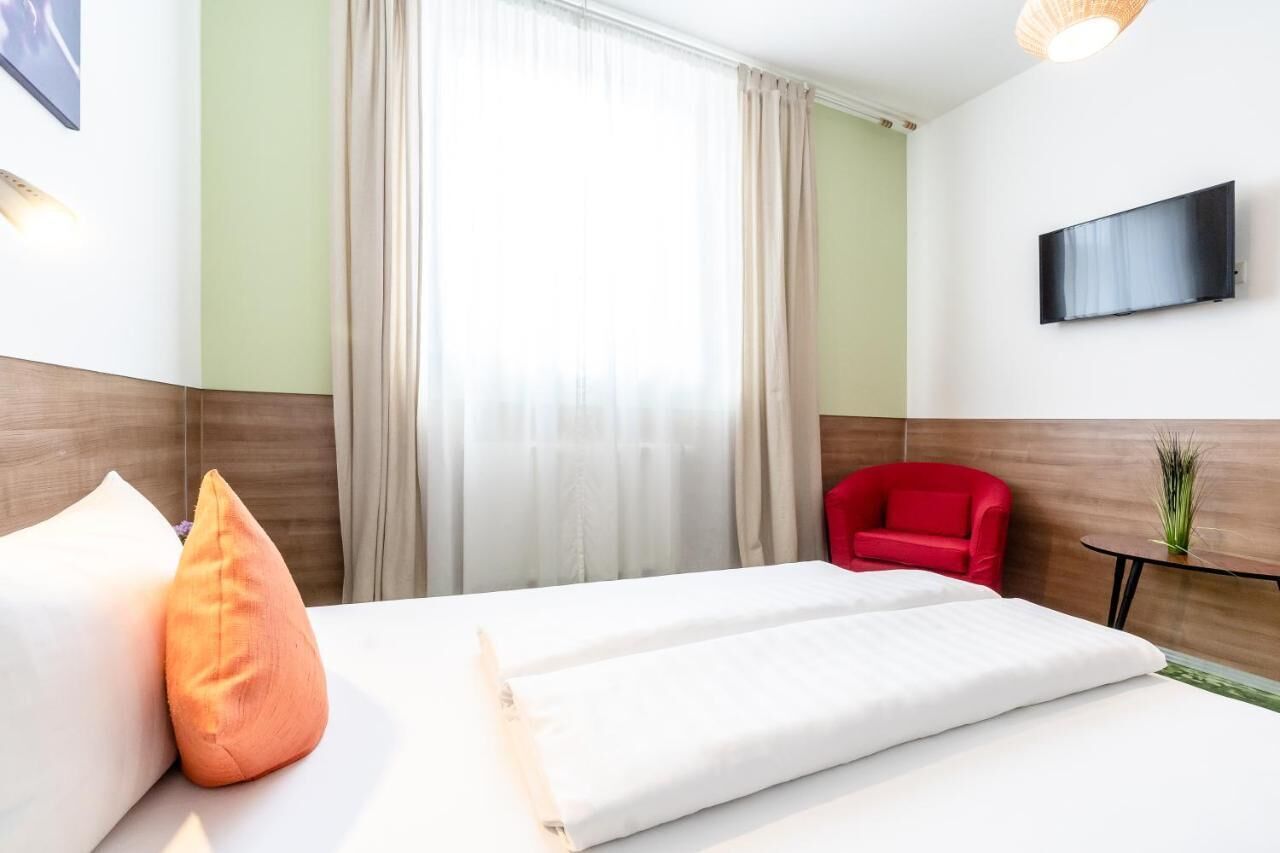 Hotellihuoneessa on sänky, punainen tuoli, pieni pöytä ja seinälle asennettu televisio.