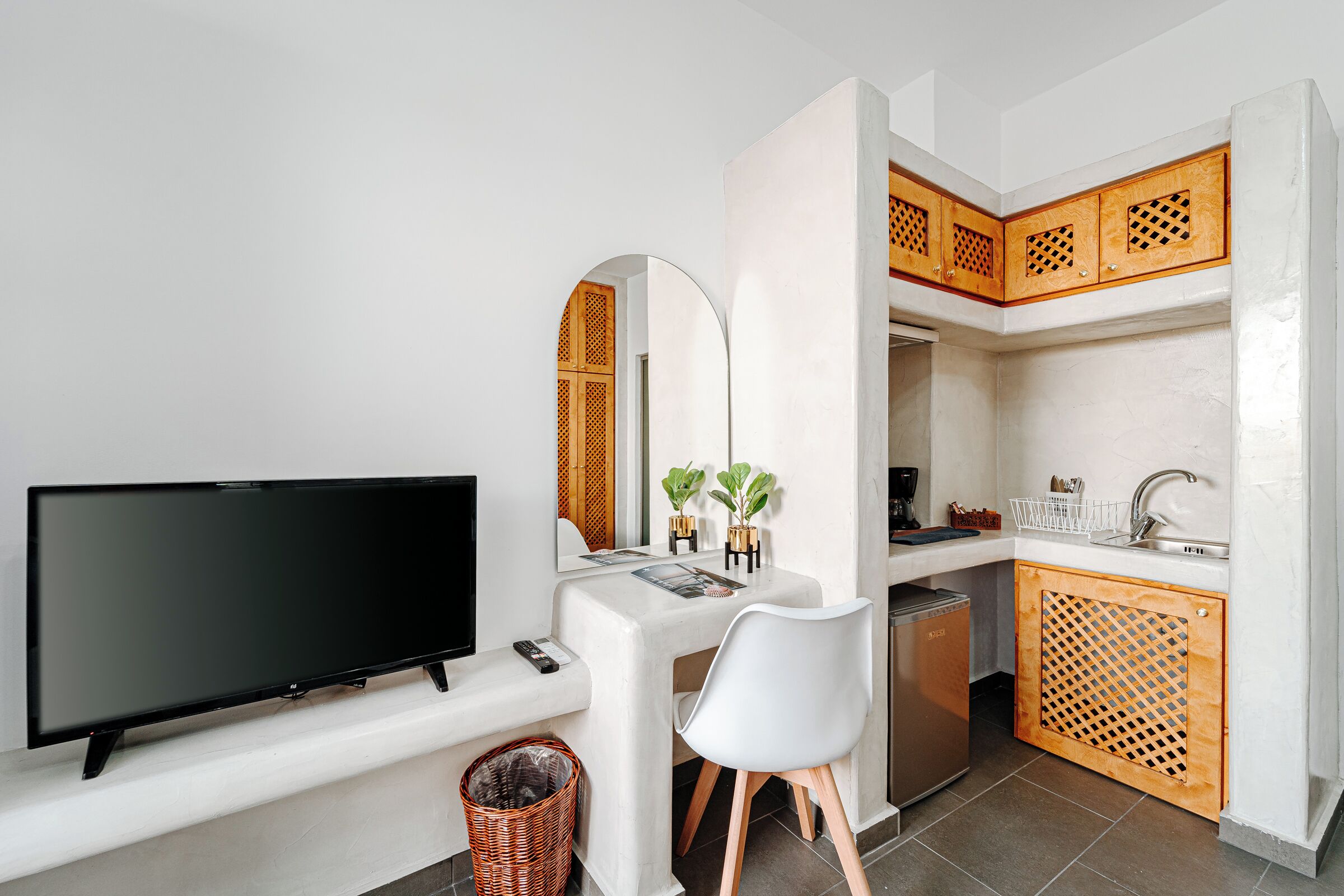 Un espacio habitable compacto con una televisión de pantalla plana, un escritorio blanco, una silla blanca, una pequeña cocina con armarios de madera y una cesta de mimbre.