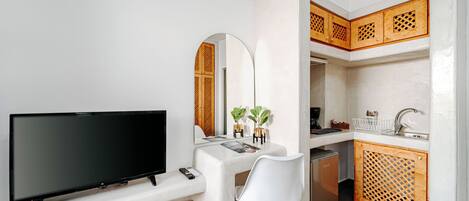 Un espacio habitable compacto con una televisión de pantalla plana, un escritorio blanco, una silla blanca, una pequeña cocina con armarios de madera y una cesta de mimbre.