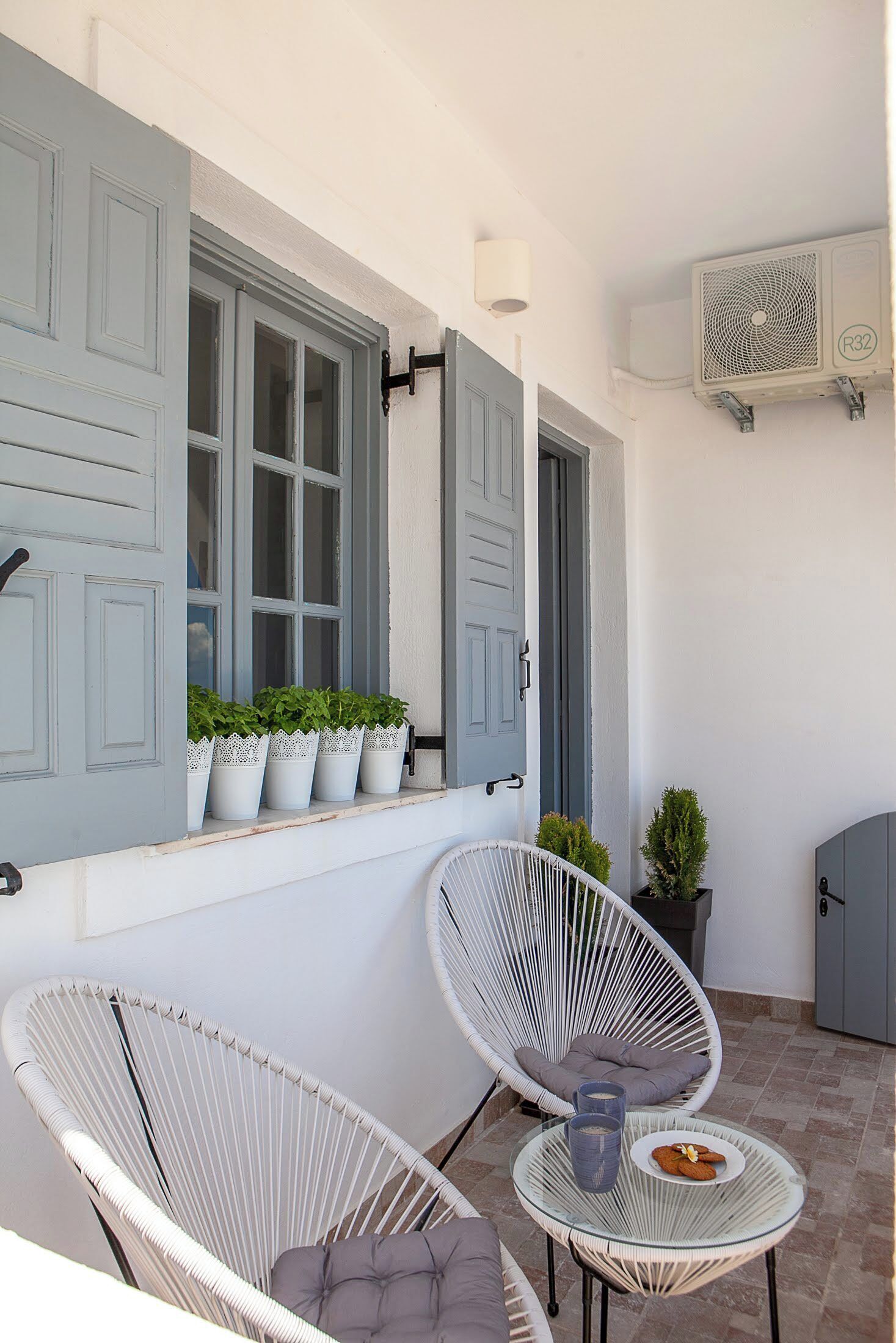 A balcony with white wicker chairs, a glass table with a plate of cookies, and potted plants.