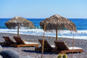 Deux fauteuils de plage en bois, surmontés de parasols en chaume, sont placés sur une plage rocheuse, face à l’océan.