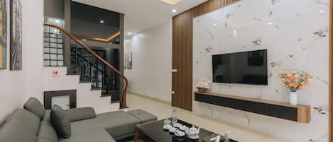 A modern living room with a sectional sofa, a coffee table, a TV mounted on a marble wall, and a staircase with a wooden railing.