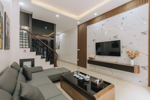 A modern living room with a sectional sofa, a coffee table, a TV mounted on a marble wall, and a staircase with a wooden railing. - Amanda Homestay - Vinhomes Ocean Park 3 (Nghia Tru)