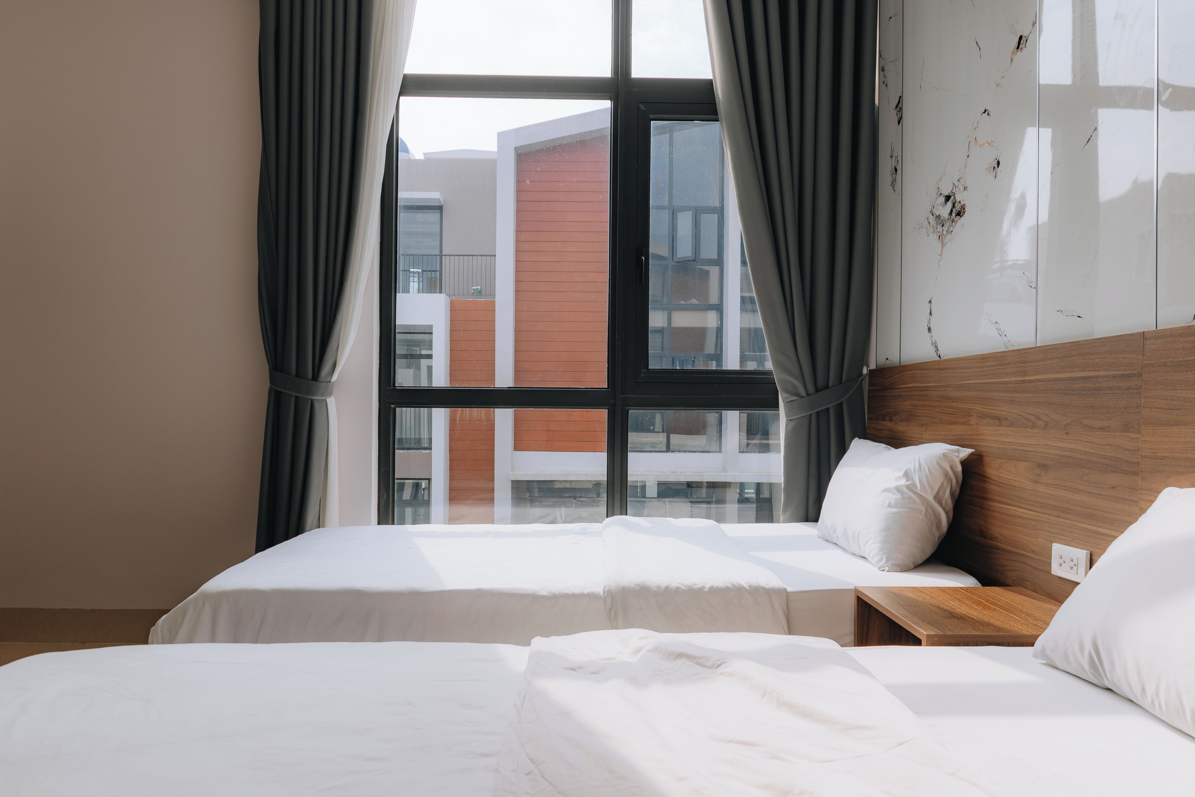 A hotel room with two beds, a wooden headboard, a window with curtains, and a view of buildings outside.