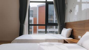 A hotel room with two beds, a wooden headboard, a window with curtains, and a view of buildings outside.