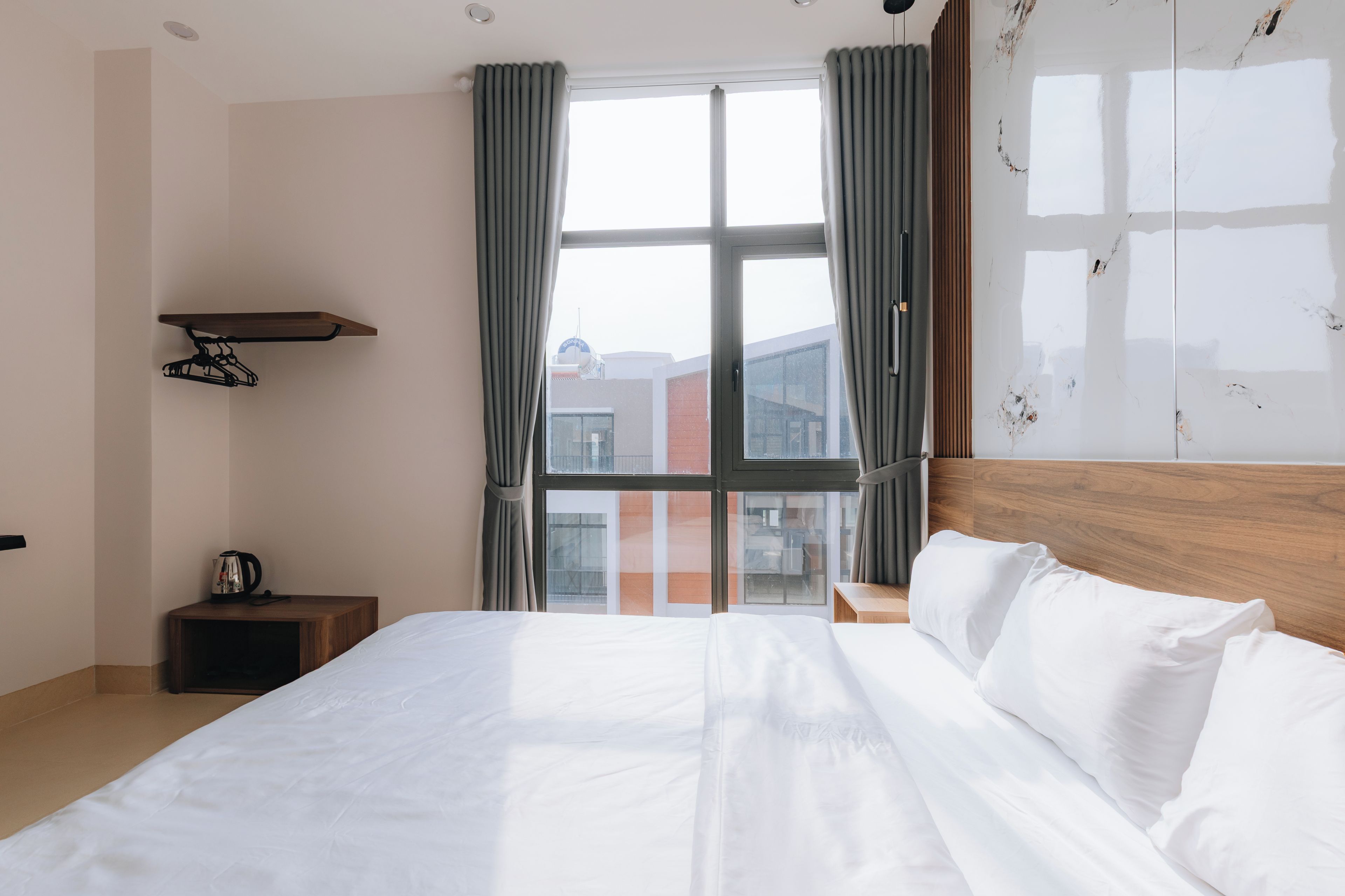 A hotel room with a large bed, a coffee maker on a side table, and a view of buildings through a window with curtains.