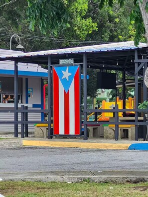 Un espacio de estar al aire libre cubierto con una bandera, un edificio con fachada azul y blanca, y un parque infantil al fondo.