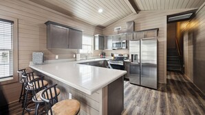 A modern kitchen with a large island, bar stools, stainless steel appliances, and a wooden floor. - Everstead Cottage - Cabin 3 (Plantersville)