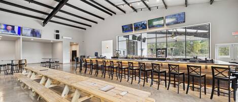 A spacious indoor area with long wooden tables and black chairs, a bar area with a counter, and large windows providing natural light.