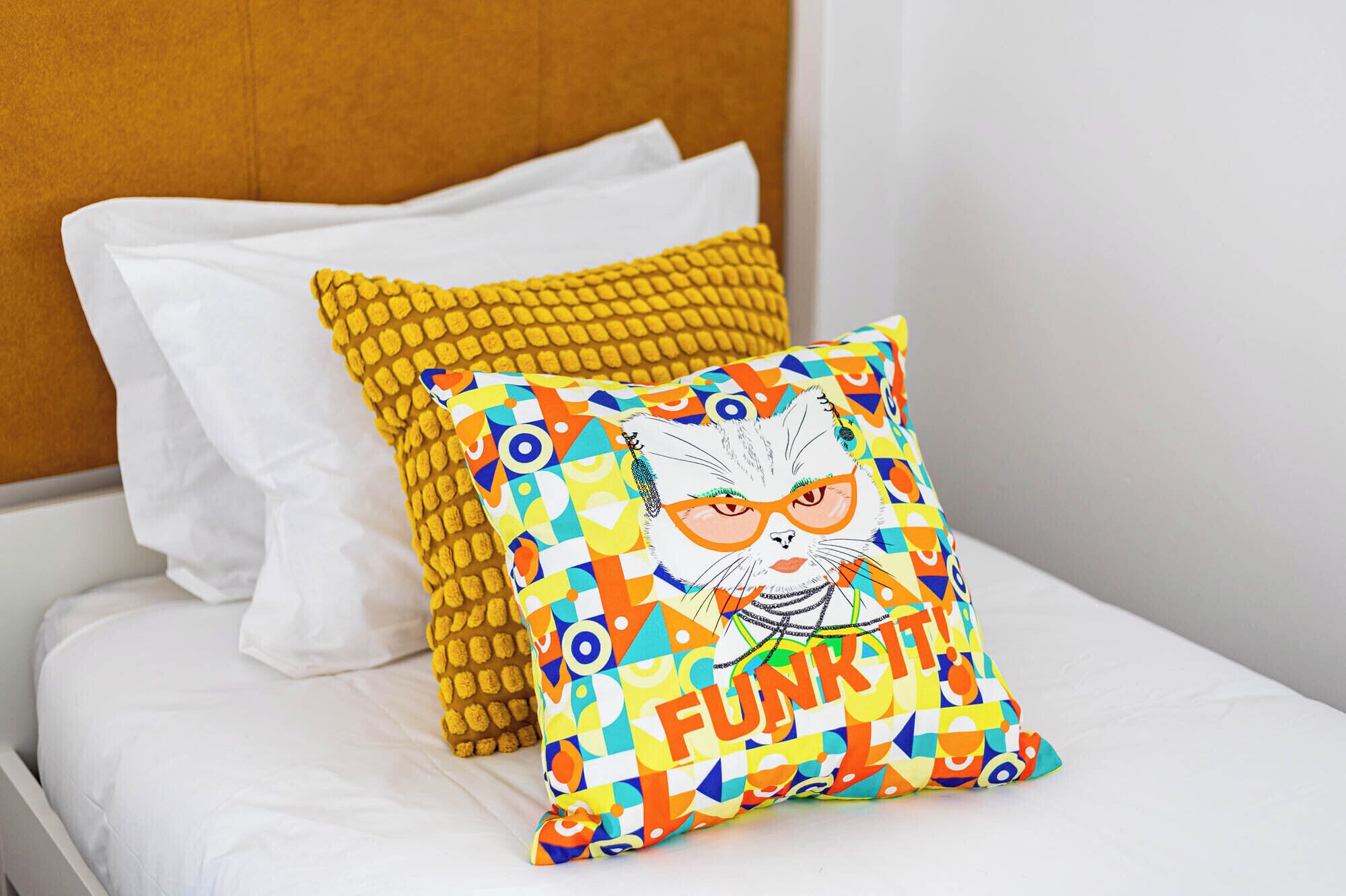 A bed with white bedding, a yellow textured pillow, and a colorful pillow with a cat illustration and the word "FUNKY".