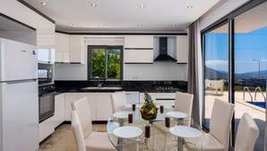 A modern kitchen with white cabinets, a glass dining table, and a view of a swimming pool. - Villa Vala in Kalkan (Kaş)