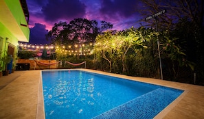 Une piscine dont le bord est revêtu de carreaux, entourée de verdure et illuminée de guirlandes lumineuses.