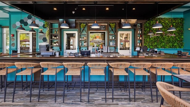 Zona de bar con mostrador, taburetes, iluminación colgante y una pared verde con un letrero.